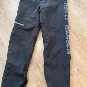 Svarta täckbyxor Helly Hansen 140 - Svarta täckbyxor från Helly Hansen med stor logga längs benet. Byxorna har elastisk midja med dragsko och meshfoder på insidan. Perfekta för kalla dagar och utomhusaktiviteter. Snygg och sportig look med reflexdetalj på benet.