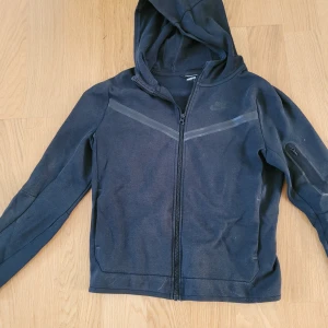 Nike Tech hoodiejacka med dragkedja - Nike tech hoodiejacka i svart med hel dragkedja framtill och huva. Den är svart I färgen, men vlivit lite urtvättad så börjar bli mer åt det grå hållet. 