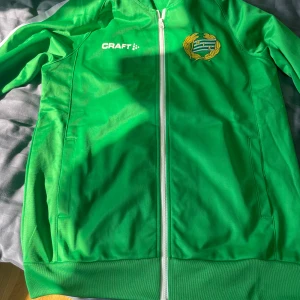 Grön Hammarby jacka från Craft S - Grön jacka från Craft med Hammarby-emblem på bröstet och stort HAMMARBY-tryck på ryggen. Jackan har vit dragkedja, ribbade muddar och två fickor. Perfekt för dig som vill representera Bajen på match eller i vardagen.