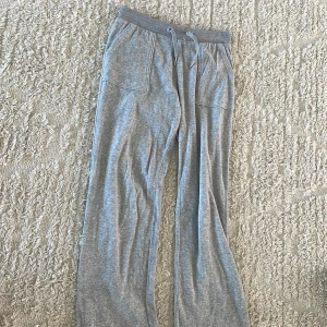 Grå mjukisbyxor Juicy Couture - Supermjuka grå mjukisbyxor från Juicy Couture med bred resår och snörning i midjan. Byxorna har raka ben, stora fickor fram och bak med lock och knappar. Perfekta för chill eller en avslappnad look.