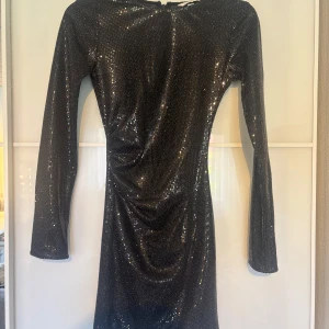 Glittrig svart klänning från H&M XS - Snygg svart kortklänning från H&M i storlek XS, täckt av glittriga paljetter. Klänningen har lång ärm, rund halsringning och en figurnära passform. Perfekt för dig som vill sticka ut med en glammig look.