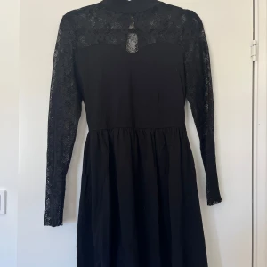 Svart klänning med spetsdetaljer - Svart klänning med långärmade spetsärmar och spets upptill. Hög hals med liten öppning fram och knappar i nacken.