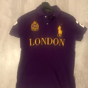 Lila Polo Ralph Lauren M - Snygg lila pikétröja från Polo Ralph Lauren med gul brodyr, stor logga och texten LONDON på bröstet. Klassisk krage, korta ärmar och broderat emblem med brittisk flagga. Nummer 2 på ryggen. Custom fit-modell i bomull.