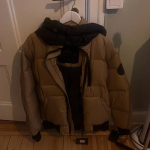 Beige dunjacka Moose Knuckles M - Säljer en oanvänd beige och svart dunjacka från Moose Knuckles i storlek M. Köpt på Pop Up Outlet på Kungsgatan 3 i vintras, säljes pga ej användning. För den som vill uppgradera sin vintergarderob! 