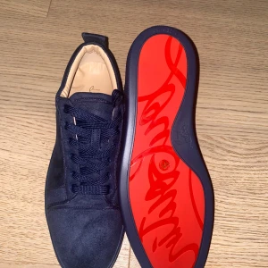 Mörkblå mocka sneakers från Louboutin - Snygga mörkblå sneakers i mocka från Christian Louboutin med klassisk röd sula och rund tå. Skorna har snörning och diskreta detaljer i samma färg för en clean look. Perfekta för dig som vill sticka ut med exklusiv stil och lyxig känsla.