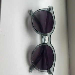 Runda solglasögon med lila glas - Snygga runda solglasögon från Glitter med transparent grå båge och mörklila glas. Perfekt accessoar för att lyfta din stil med en cool retrovibe. Lätta och bekväma att bära hela dagen.