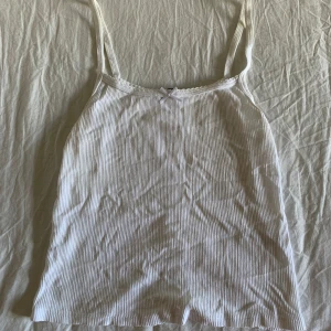 Vit ribbad linnetopp Brandy Melville - Supersöt vit ribbad linnetopp från Brandy Melville med tunna axelband och liten rosett framtill. Toppen har en smal passform och är perfekt för varma dagar. Diskret spetskant upptill ger en extra gullig touch.