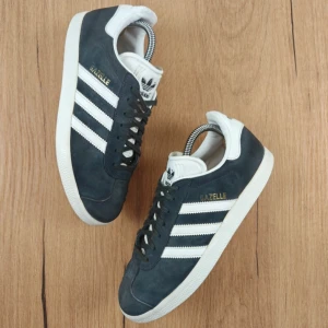 Adidas Gazelle sneakers i mörkblå mocka - Adidas Gazelle sneakers i mörkblå mocka med klassiska vita ränder och vit sula. Skorna har rund tå, snörning och vit hälkappa med Adidas-logga. Perfekt för dig som gillar retrostil och streetwear.