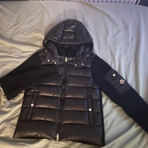 Svart Moncler dunjacka  - Snygg svart dunjacka från Moncler med stickade ärmar och vadderad kropp. Jackan har huva, dragkedja framtill och två fickor med tryckknappar. Moncler-logga på ena ärmen ger en exklusiv touch. Perfekt för kyliga dagar när du vill hålla stilen.