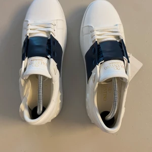 Valentino Open  - Snygga Valentino Garavani Open sneakers i vitt skinn med bred marinblå rem över vristen. Klassisk rund tå, vita snören och cool mönstrad sula. Kommer med originalkartong och dustbag. Perfekta för dig som vill ha en stilren men lyxig look.