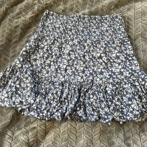 Blommig volangkjol från 157, blå/vit - Supersöt kort kjol från 157 i blått med vitt blommönster. Kjolen har smock i midjan för skön passform och volang nertill som ger en flowy vibe. Perfekt för dig som gillar en feminin och avslappnad stil.