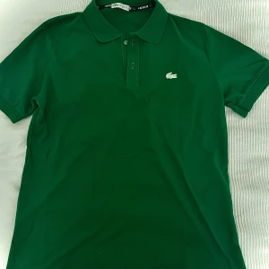 Grön pikétröja från Lacoste M - Snygg grön pikétröja från Lacoste i klassisk modell med korta ärmar och krage. Tröjan har knappar framtill och den ikoniska krokodilloggan på bröstet. Tillverkad i mjuk bomull som andas, perfekt för en clean och sportig look.
