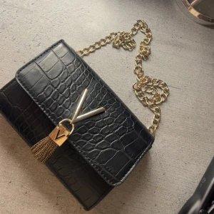 Mario Valentino Svart Axelremväska  - Snygg svart axelväska från Valentino med krokopräglat mönster och guldfärgad kedja. Väskan har ett stort V i metall framtill och dekorativa guldkedjor som detalj. Perfekt storlek för det viktigaste och ger en lyxig känsla till din outfit.💕skriv gärna om ni har fler frågor!!!