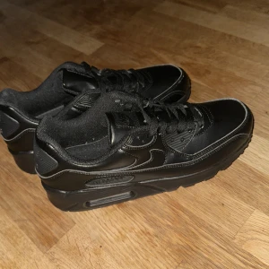 Svarta Nike Air Max sneakers - Snygga helsvarta Nike Air Max 90 sneakers med klassisk siluett och synlig luftbubbla i sulan. Skorna har rund tå, snörning och är tillverkade i skinn och syntet. Perfekta för dig som gillar en clean och sportig look. Har använts ungefär 2 gånger max.