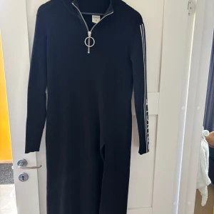 Svart långklänning Pringle x H&M - Svart ribbad långklänning med lång ärm från Pringle of Scotland x H&M. Klänningen har en stor silverfärgad dragkedja framtill och vita ränder med logga längs ärmen. 