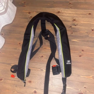 Svart flytväst Baltic Winner - Säljer en svart flytväst Baltic Winner med grå och gula detaljer. Västen har justerbara remmar och spänne framtill samt reflexdetaljer. Perfekt för båtliv och vattenaktiviteter. Snygg och funktionell design med tydlig Baltic-logga på sidan. Nyköpt 2200kr och använd 2 gånger 