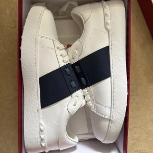 Valentino Garavani - Snygga Valentino Garavani Open sneakers i vitt skinn med bred marinblå rem över mitten. Klassisk rund tå, vita snören och coola nitar på hälen. Perfekta för dig som gillar stilrena och exklusiva sneakers med en twist.