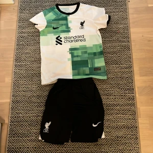 Liverpool Salah bortatröja Nike set - Liverpool FC bortatröja med M. Salah 11 på ryggen. Tröjan är vit med gröna grafiska mönster och svarta detaljer, tillverkad i lätt och ventilerande material. Matchande svarta shorts med LFC-logga och Nike swoosh. Perfekt för dig som älskar fotboll och Salah! Lite slitig text där bak