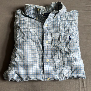 Rutig skjorta från Ralph Lauren  - Snygg rutig skjorta från Ralph Lauren i blått och vitt. Slim fit-modell i 100% bomull med stretch. Klassisk krage, knappar framtill och broderad logga på bröstet. Perfekt för dig som gillar stilrena och tidlösa plagg. Hör av er vid minsta lilla fråga 