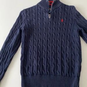Marinblå stickad tröja Polo Ralph Lauren - Snygg marinblå kabelstickad tröja från Polo Ralph Lauren med halv dragkedja och röd broderad logga på bröstet. Tröjan har hög krage, långa ärmar och ribbade muddar. Perfekt för dig som gillar klassisk och stilren look. Färgen är mörkblå skick 8.5/10 använd fåtal gånger NYTT PRIS 1799