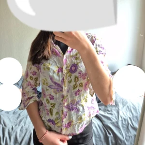 Blommig skjorta - Säljer en tunn och luftig skjorta med blommigt mönster i lila, gult och grönt på vit botten. Skjortan har trekvartsärmar med lätt volang vid ärmslut och knäppning framtill. Perfekt för dig som gillar färg och vill sticka ut lite extra.