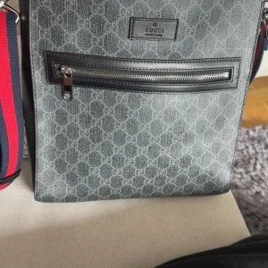 Gucci axelväska med GG-mönster - Snygg axelväska från Gucci i grått med klassiskt GG-mönster över hela väskan. Väskan har en bred, justerbar axelrem i rött och marinblått, samt detaljer i svart skinn. Framsidan har en praktisk dragkedjeficka och silverfärgade metalldetaljer.