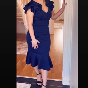 Marinblå volangklänning med v-ringning - Snygg marinblå klänning med v-ringning och volangdetaljer på axlarna. Klänningen är figurnära med volangkant nertill och har korta ärmar. Perfekt för dig som vill sticka ut med en elegant och modern look.