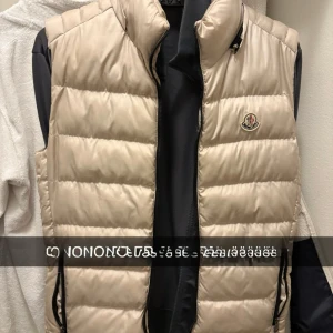 Beige dunväst från Moncler - Snygg beige dunväst från Moncler med quiltad design och hög krage. Västen har dragkedja framtill och två fickor med dragkedja. Klassisk Moncler-logga på bröstet. Perfekt att slänga över en hoodie för en clean look.