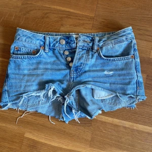 Blå jeansshorts - Snygga blå jeansshorts från lager 157 med låg midja, slitna detaljer och fransig kant. Klassisk femficksmodell med knappar framtill och råa kanter för en cool, avslappnad vibe. Perfekta för varma dagar och festivalhäng. Står ingen storlek men jag skulle säga ungefär XS/32