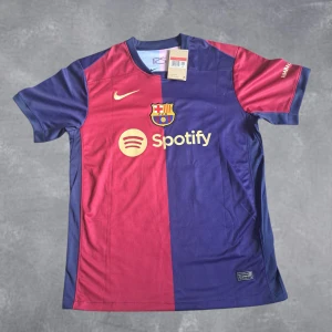 FC Barcelona fotbollströja  - Säljer en officiell FC Barcelona fotbollströja från Nike med Raphinha 11 på ryggen. Tröjan är kortärmad och har klassisk halv-halv design i vinröd och marinblå med klubbmärke, sponsorer och gula detaljer. Perfekt för dig som älskar Barca och vill sticka ut på läktaren.