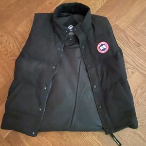 Svart dunväst från Canada Goose - Säljer en svart dunväst från Canada Goose med klassisk logga på bröstet. Västen har hög krage, tryckknappar och dragkedja framtill samt två sidofickor. Perfekt för lager-på-lager och håller dig varm under kyliga dagar.