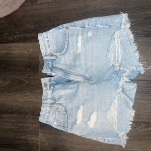 Ljusblåa jeansshorts med slitningar - Snygga ljusblå jeansshorts med hög midja och råa, fransiga kanter. Shortsen har flera slitningar framtill för en trendig, avslappnad look. 