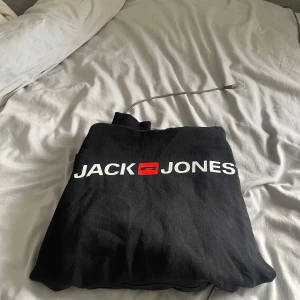 Svart hoodie från Jack & Jones - Svart hoodie från Jack & Jones med stor vit logga och röd detalj på bröstet. Klassisk modell med huva och snörning, perfekt för en chill och avslappnad stil. Mjuk bomullskänsla och lång ärm.