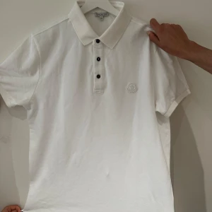 Vit pikétröja från Moncler - Säljer en stilren vit pikétröja från Moncler med klassisk krage och tre svarta knappar framtill. Tröjan har broderad Moncler-logga på bröstet och är gjord i mjuk bomull. Perfekt för dig som gillar en clean och exklusiv look.