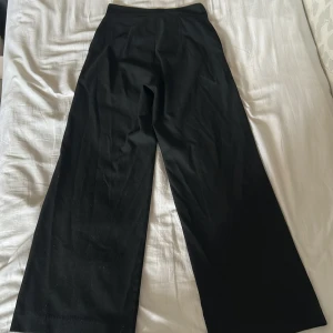 Svarta kostymbyxor från Lindex - Svarta kostymbyxor från Lindex i modellen Lykke. Byxorna har hög midja och vida ben för en snygg och trendig siluett. Perfekta att styla med sneakers eller boots. Materialet är mjukt och följsamt, vilket gör dem riktigt bekväma.