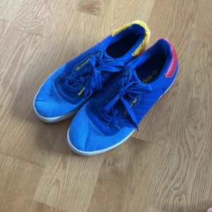 Adidas blå sneakers med färgglada detaljer - Unika Adidas sneakers i blått med snörning, vit sula och coola detaljer: röd häl på ena skon och gul frotté på den andra. Skorna har klassisk Adidas-logga i gult och är tillverkade i textil och syntet. Perfekta för dig som vill sticka ut med färg och stil.