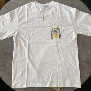 Vit t-shirt från Casablanca med tryck - Vit t-shirt från Casablanca med färgglatt tryck av en byggnad och gröna blad på bröstet och ryggen. Klassisk passform och rund halsringning. Perfekt för dig som gillar stilrena plagg med unika detaljer. Perfekt till sommaren