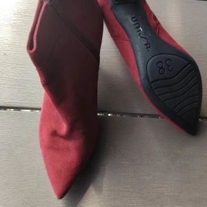 Vinröda stövletter från Unisa - Snygga vinröda stövletter från Unisa i mjuk mocka. Skorna har spetsig tå, låg klack och dragkedja på sidan för enkel påtagning. Perfekta för att lyfta din outfit med en trendig touch.