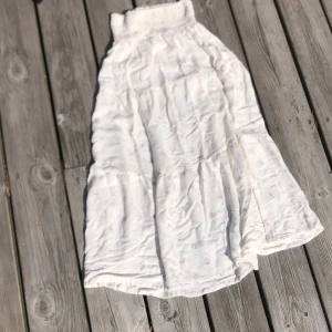 Vit långkjol med blommigt mönster - Supersöt vit långkjol från 157 med små rosa och blå blommor. Kjolen har en bred, smockad midja som är riktigt bekväm och ger en snygg passform. Materialet känns mjukt och luftigt, perfekt för varma dagar. Den har volanger som ger extra flow och en romantisk vibe.