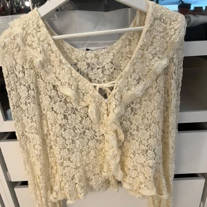 Spetsblus med volang från Gina Tricot - Säljer en romantisk spetsblus från Gina Tricot i storlek S. Blusen är krämvit med blommigt spetsmönster, volangdetaljer längs halsringningen och framtill samt snörning vid bröstet. Långärmad och lätt transparent, perfekt för dig som gillar vintagevibe och detaljer. Aldrig använd! 