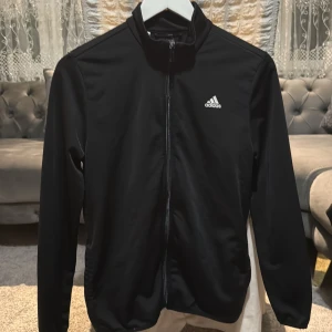 Svart Adidas tröja - Svart vit tröja från Adidas med vit logga på bröstet och stor logga på ryggen. Jackan har hel dragkedja framtill, ståkrage och långa ärmar. Tillverkad i Primegreen-material som är syntetiskt och miljövänligt. Perfekt för sportiga dagar och chill.