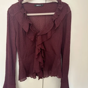 Vinröd blus med volanger Gina Tricot - Supersnygg vinröd blus från Gina Tricot med volanger längs v-ringningen och framtill. Lätt transparent material och långärmad modell som sitter fint och ger en feminin vibe. Perfekt att styla med jeans eller kjol för en trendig look.