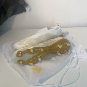 Adidas F50 fotbollsskor i vitt och guld - Säljer ett par Adidas F50 fotbollsskor i vitt med snygga gulddetaljer och guldplatta under sulan. Skorna har en strömlinjeformad design utan snörning och är tillverkade i syntetmaterial för lätt känsla på planen. Perfekt för dig som vill sticka ut med stil på matchen.