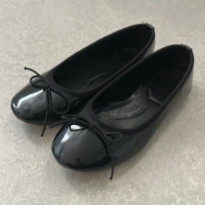 Svarta ballerinaskor med rosett - Snygga svarta ballerinaskor i glansigt syntetmaterial med rund tå och en söt rosett framtill. Skorna har en klassisk och stilren look som passar perfekt till jeans eller klänning. Platt sula för skön komfort hela dagen. Använt bara en gång