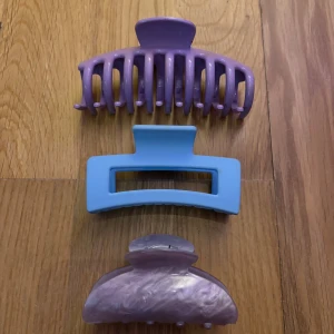 Tre hårklämmor i lila och blå - Säljer ett set med tre hårklämmor i olika former och färger. En lila klassisk klämma med rundade taggar, en blå rektangulär klämma med matt finish och en mindre lila klämma med skimrande marmor-effekt. Perfekt för att fixa håret snabbt och snyggt.