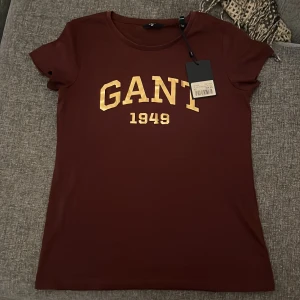 Vinröd GANT t-shirt med guldtryck - Snygg vinröd t-shirt från GANT i storlek XS med korta ärmar och rund hals. Framsidan har ett stort guldigt tryck med GANT 1949. Materialet är mjuk bomull som känns skönt mot huden. Perfekt för dig som gillar klassisk college-stil med en modern twist.