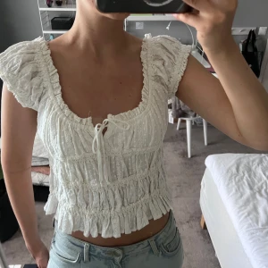 Vit blus med volanger och knyt - Supersöt vit blus med volanger och smockad midja. Blusen har korta puffärmar som kan bäras både på axeln och offshoulder, samt en U-ringad hals med knyt framtill. 