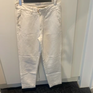 Vita/beiga linnebyxor från These Glory Days - Snygga vita linnebyxor från These Glory Days i storlek 46. Byxorna har en klassisk rak passform med fickor fram och bak, bälteshällor och knappstängning. Materialet är 52% linne och 48% bomull, vilket gör dem både luftiga och stilrena.