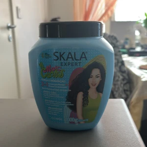 Skala Expert Mais Cachos hårkräm - Stor burk med Skala Expert Mais Cachos 2-i-1 hårkräm för lockigt hår. Kommer i en ljusblå plastförpackning med svart lock och färgglad etikett. Innehåller 1000g krämig, vit produkt som används som leave-in eller balsam. Perfekt för dig som vill ha definierade och återfuktade lockar.