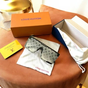 Louis Vuitton monogram solglasögon - Säljer ett par svarta Louis Vuitton solglasögon med bred, rektangulär sköldform och ikoniskt monogrammönster över hela linsen. Bågarna är svarta med guldfärgade LV-detaljer på sidorna. Glasen är tonade i grått och har en exklusiv vibe, perfekt för dig som gillar statement-accessoarer.